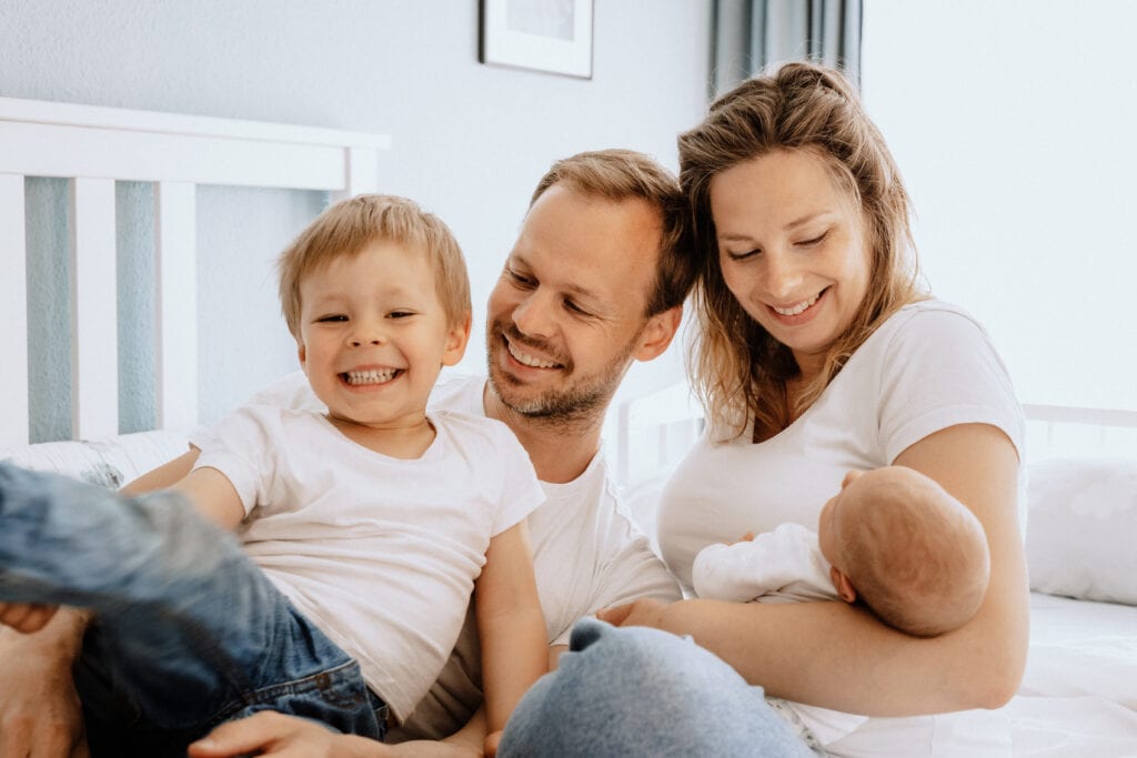 Eine vierköpfige Familie sitzt zusammen auf einem Bett. Die Mutter hält lächelnd ein Baby im Arm. Der neben ihr sitzende Vater schaut auf das ältere Kind, das lacht und in Bewegung ist. Alle tragen lässige weiße Oberteile und Jeans. Ein schönes Beispiel von Familienfotografie.