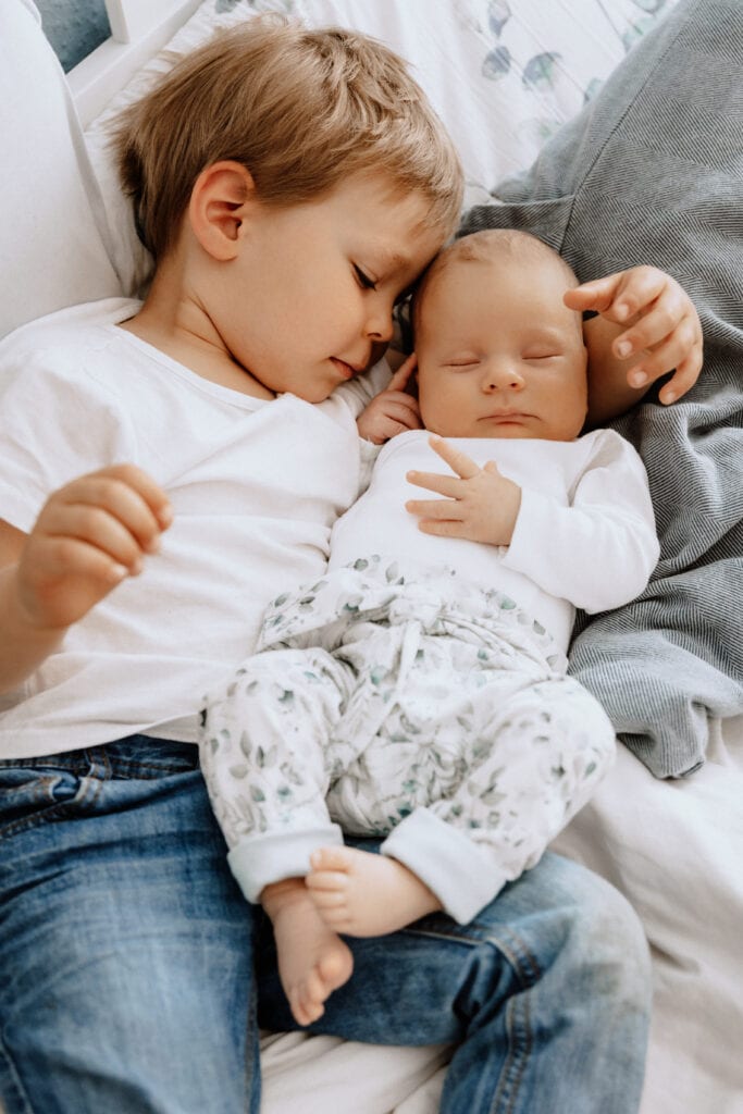 Ein kleines Kind und ein Baby liegen eng beieinander auf einem Bett. Das ältere Kind, das ein weißes Hemd und Jeans trägt, hält das Baby, das ein weißes Oberteil und gemusterte Hosen trägt, sanft im Arm. Beide Kinder wirken entspannt und ruhig.