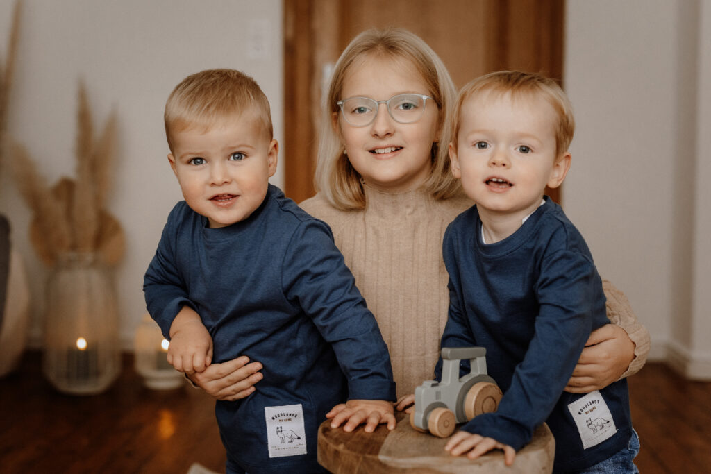 Ein junges Mädchen mit Brille und beigem Pullover lächelt, während es zwei Kleinkinder in passenden blauen Hemden hält. Sie sind drinnen, mit einem Holzspielzeug auf einem kleinen Hocker vor ihnen und verschwommenen Dekorationen im Hintergrund – perfekt eingefangen von einem Familienfotografen aus Havixbeck im Münsterland.