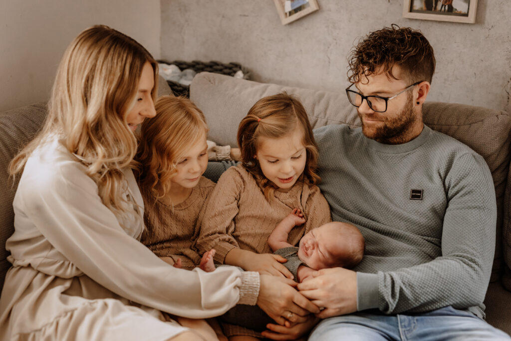 Eine Familie sitzt gemütlich auf der Couch, liebevoll festgehalten von einer Familienfotografin aus Havixbeck. Eine Frau wiegt ein Baby, während zwei Mädchen ihre Geschwister anstarren, während der Mann und die Frau herzlich lächeln und sich ganz ihren Kindern widmen.