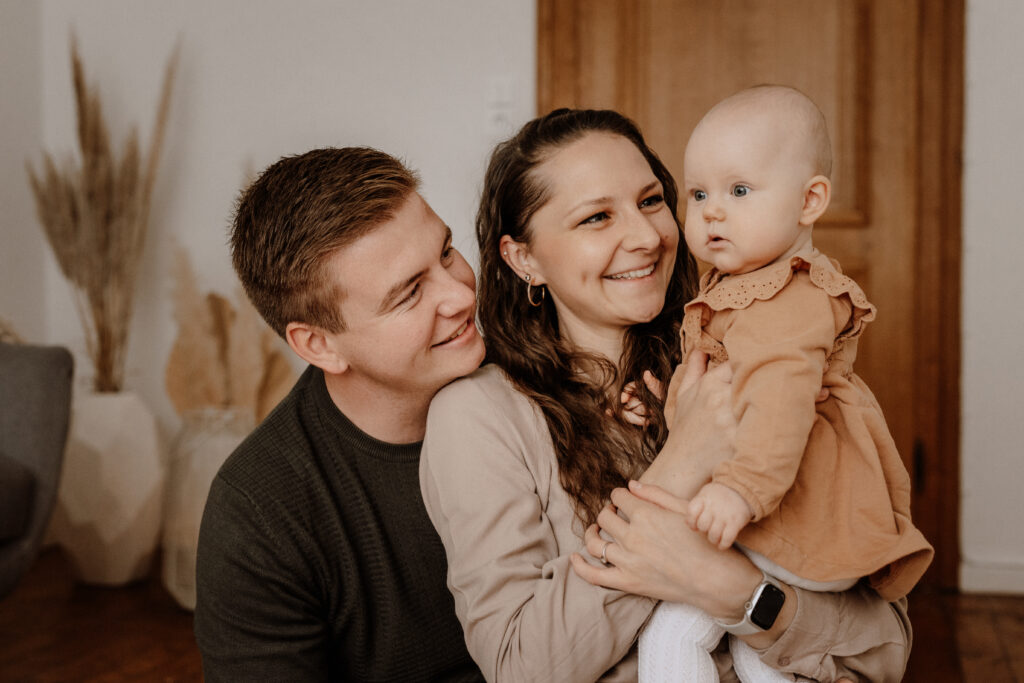 Ein Paar sitzt eng beieinander und lächelt sich an, während die Frau ein Baby hält, was an ein kürzliches Babybauch-Shooting erinnert. Das Baby, das ein braunes Outfit trägt, schaut zur Seite. Sie befinden sich in einem warm beleuchteten Raum mit Holzakzenten und getrockneten Pflanzen.