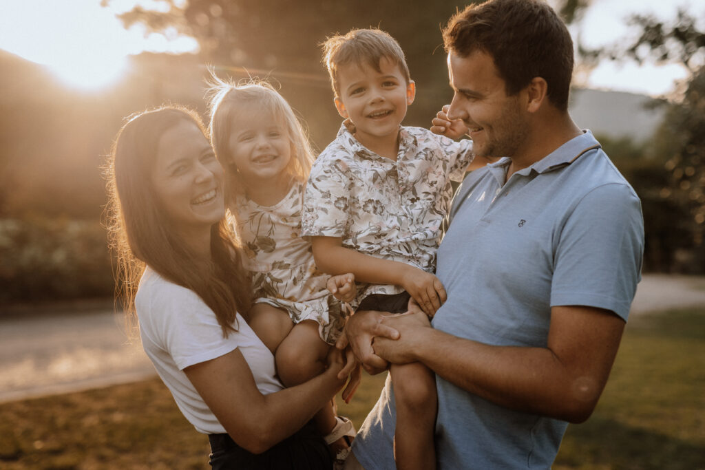 Eine lächelnde vierköpfige Familie genießt einen sonnigen Tag im Freien, perfekt eingefangen von einer Familienfotografin aus Billerbeck. Die Eltern in legerer Kleidung halten ihre beiden kleinen Kinder in gemusterten Hemden, während das helle Sonnenlicht eine warme, strahlende Atmosphäre schafft.